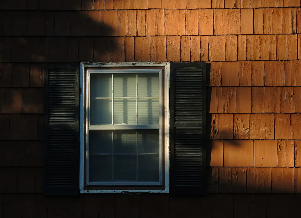 Dark louvered shutters flanking a white-framed window on weathered wood shingle siding in evening light