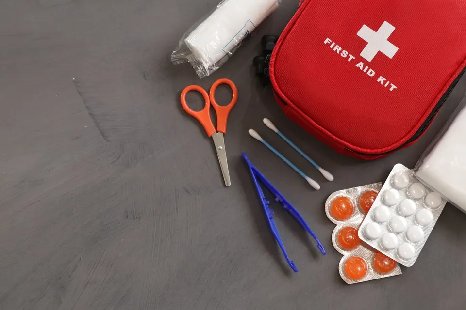 Red first aid kit pouch beside gauze, scissors, tweezers, cotton swabs and blister packs of tablets