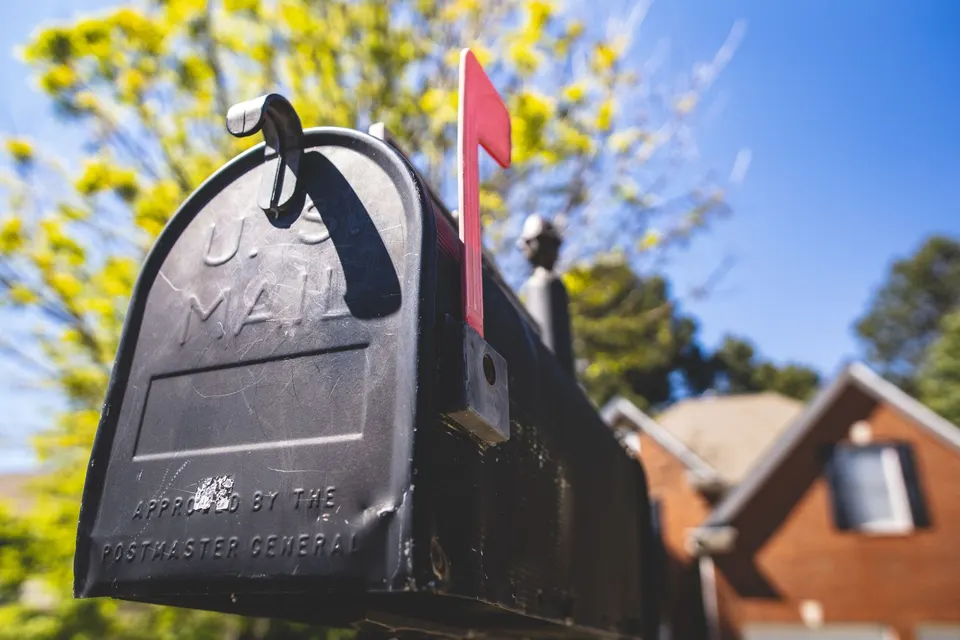 Closed black curbside mailbox with its red flag raised, a brick house blurred behind it