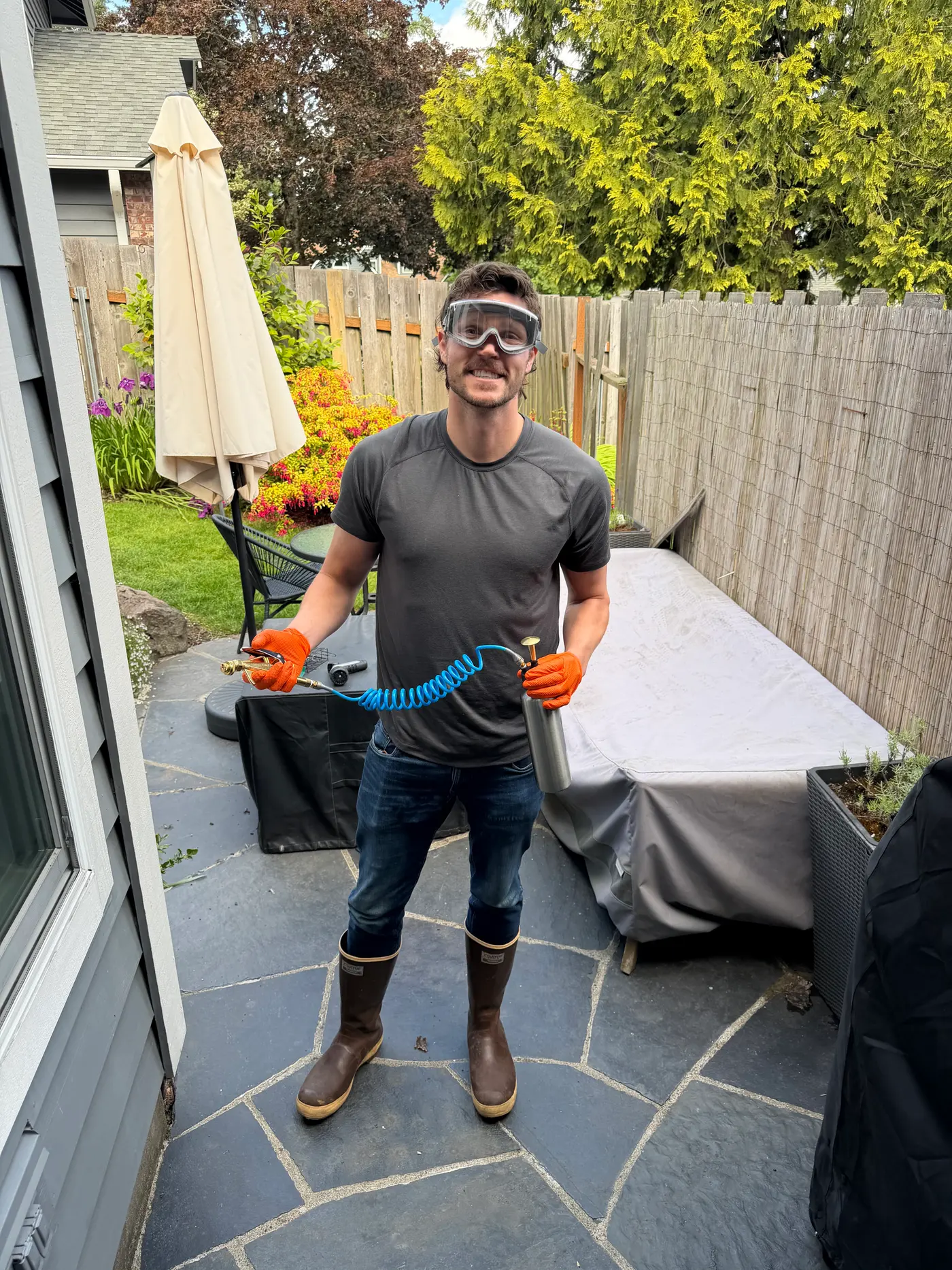 Sam in safety goggles and orange gloves holding a handheld pressurized sprayer on a backyard patio