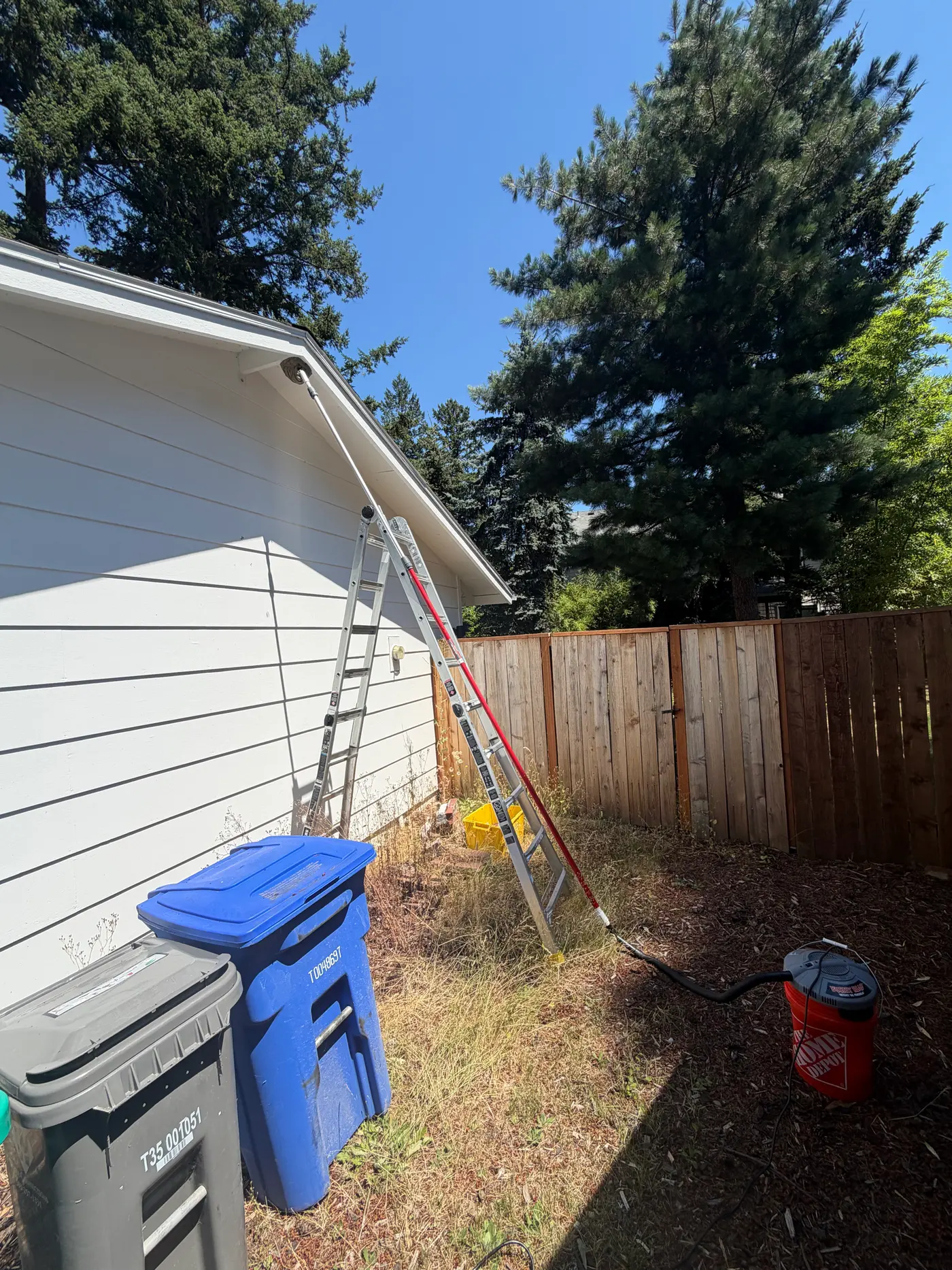 Extension ladder and long telescoping pole reaching a house eave, hose running to a bucket vacuum