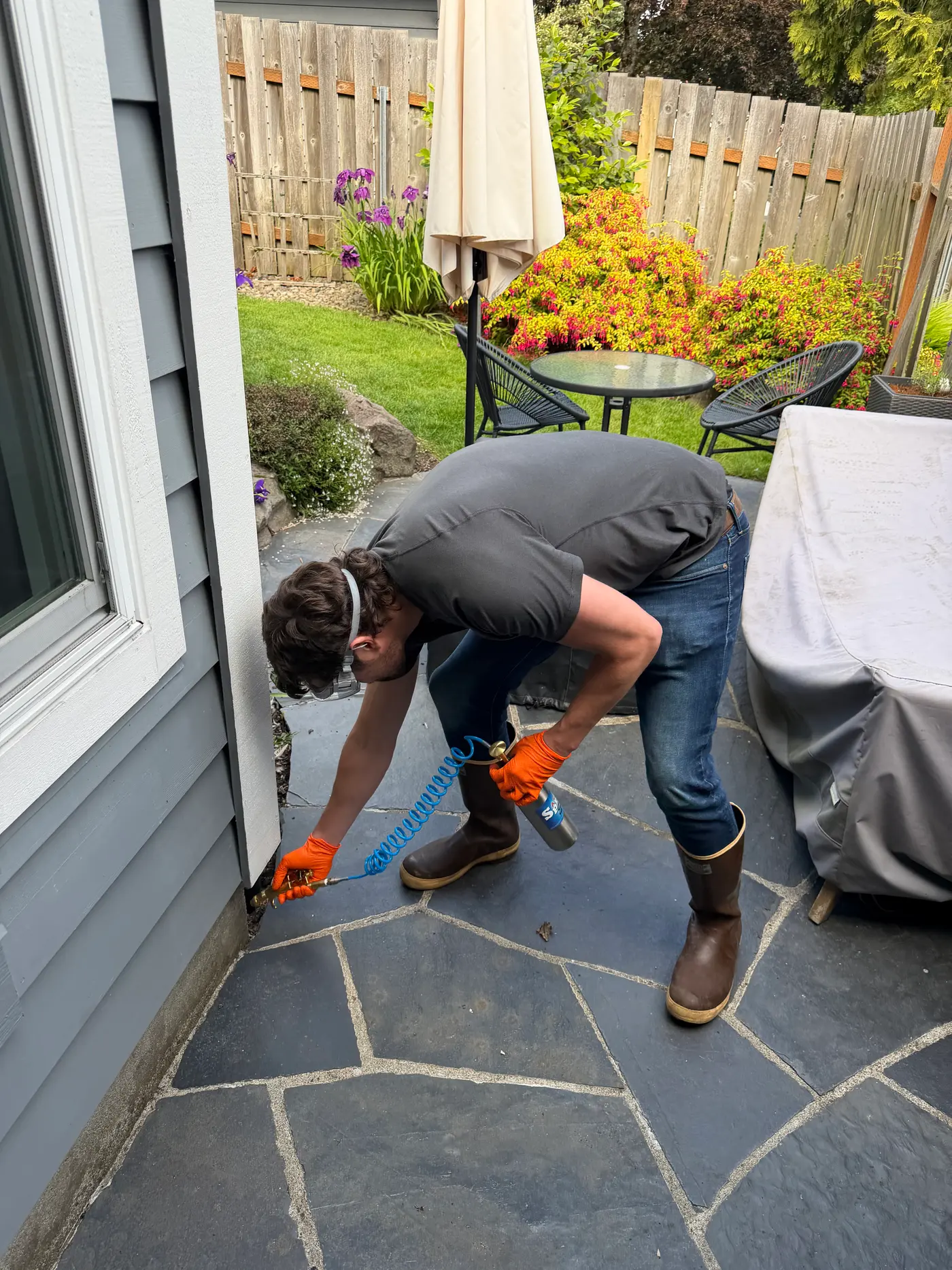 Sam in orange gloves spraying pest treatment into the base of house siding beside a backyard patio