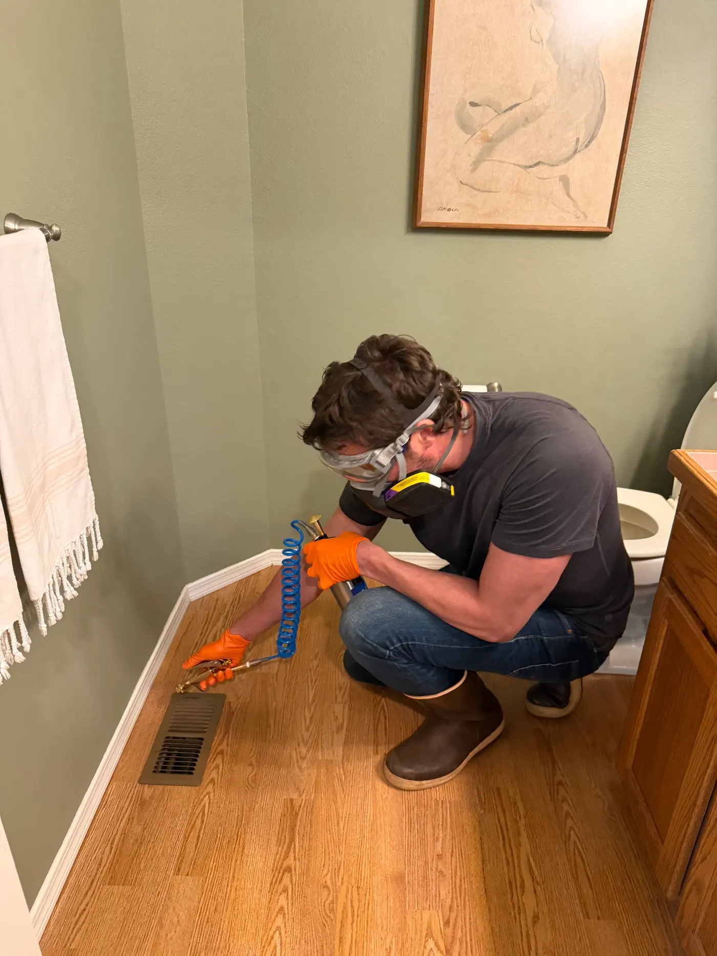 Sam in a respirator and goggles applying pest control treatment into a bathroom floor vent register
