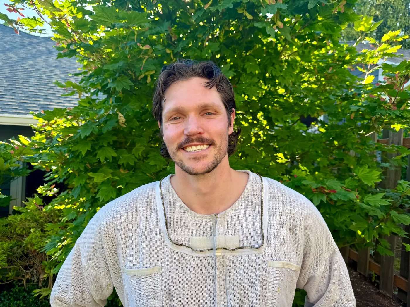 Sam smiling in a white bee suit standing outdoors in a green residential yard