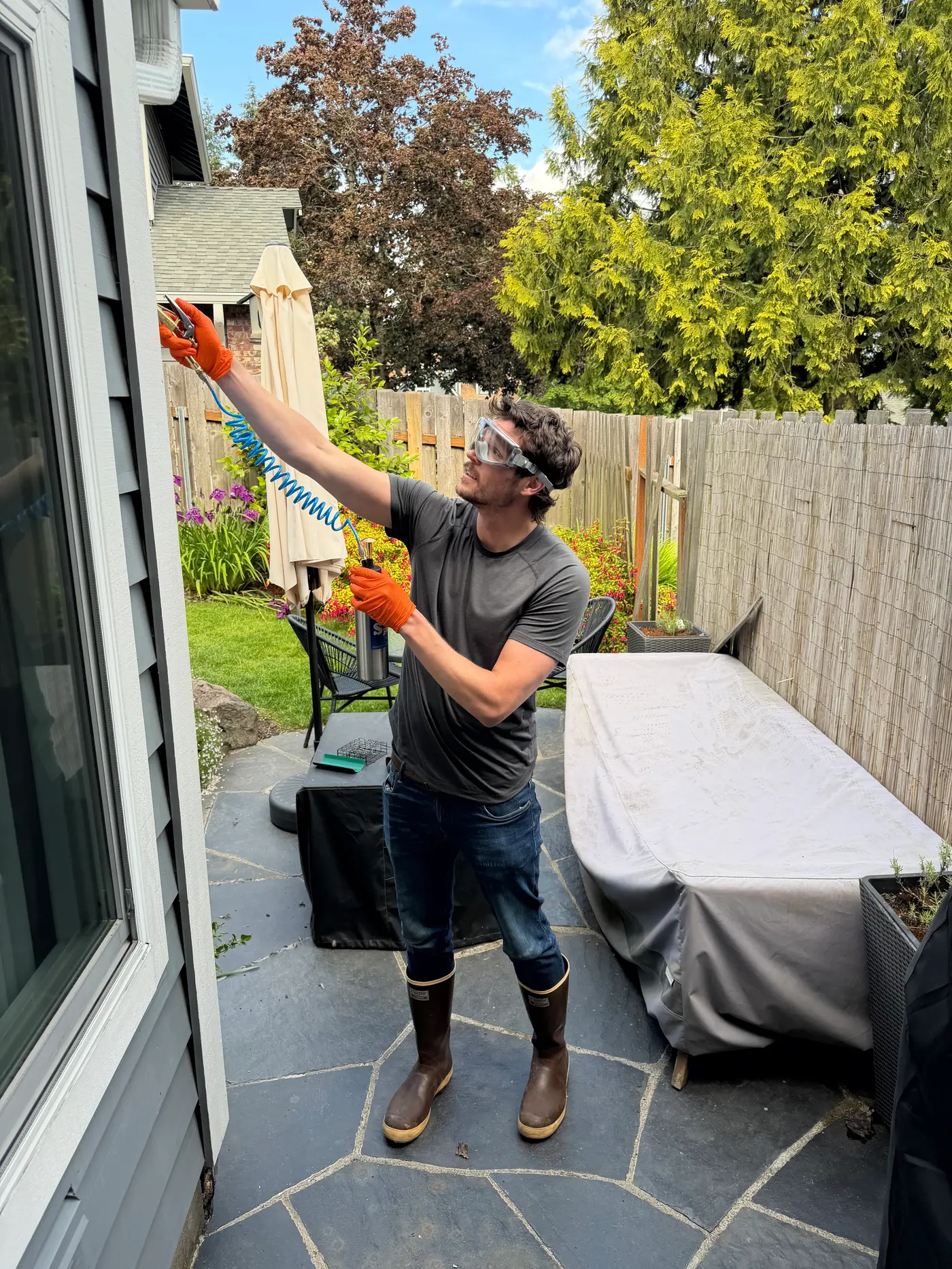 Sam in safety goggles and orange gloves reaching a hand tool up under a house eave