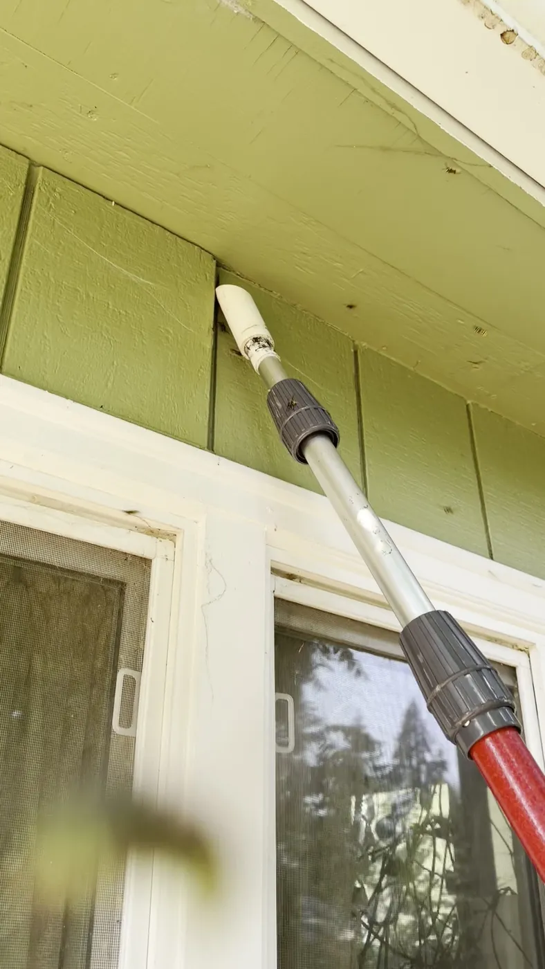 White dust applicator tip on an aluminium extension pole held against the underside of a soffit while treating a yellow jacket nest