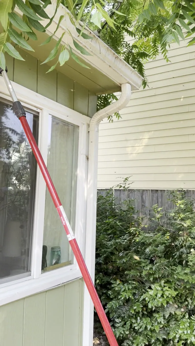 Green board-and-batten bay window and cream lap siding on a Milwaukie home, with a red telescoping pole raised toward the eave during a yellow jacket treatment